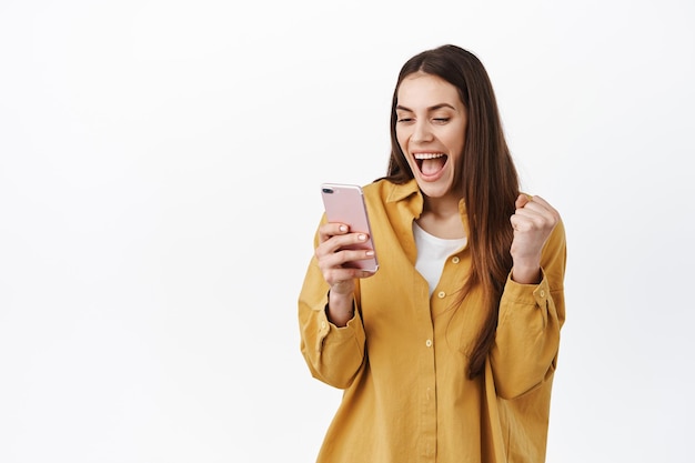 Obraz Dziewczyny świętującej Wygraną Online, Patrzącej Na Smartfona I Cieszącej Się, Wygrywającej Lub Osiągającej Codzienny Cel W Aplikacji, Czytającej Wspaniałe Wiadomości Na Ekranie Telefonu, Stojącej Na Białym Tle