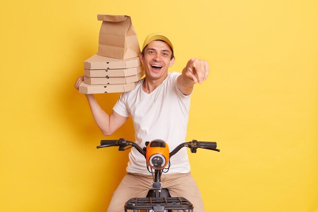 Obraz kuriera dostarcza smaczną, świeżo upieczoną pizzę, życzy ci dobrego apetytu, trzyma kartonowe pudełko w białej koszulce siedzi na szybkim motocyklu, wskazując na kamerę odizolowaną na żółtej ścianie