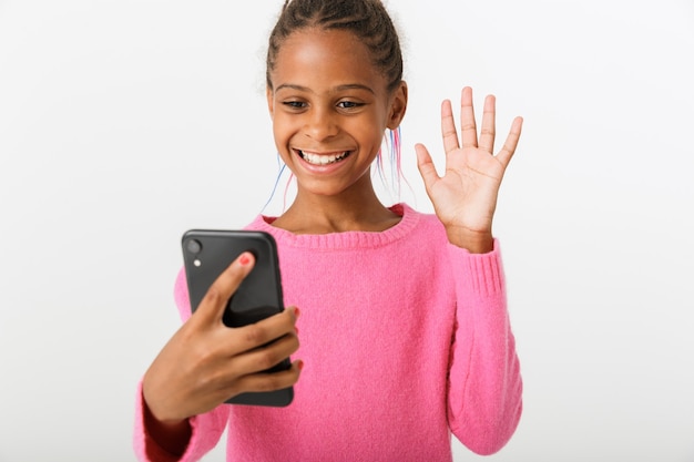 Obraz ładnej afroamerykańskiej dziewczyny trzymającej telefon komórkowy i machającej ręką na białym tle nad białą ścianą
