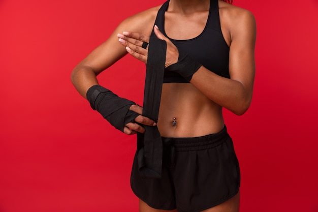 Obraz Lekkoatletycznego African American Kobieta W Czarnej Odzieży Sportowej Noszących Bandaże Sportowe Na Rękach, Odizolowane Na Czerwonej ścianie