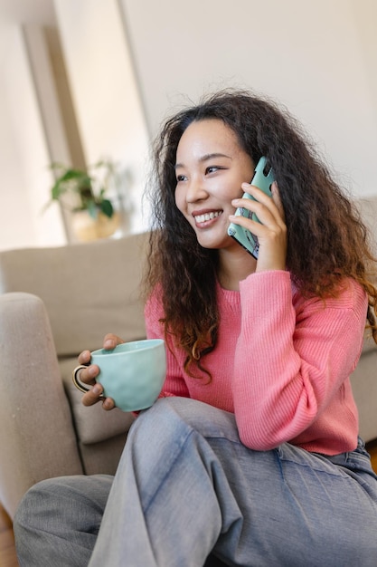 Obraz Miłej Azjatyckiej Młodej Kobiety Używającej Telefonu Komórkowego I Rozmawiającej Siedząc Na Kanapie I Pijąc Kawę W Salonie