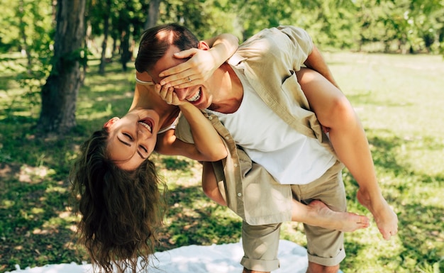 Obraz młodego mężczyzny niosącego swoją piękną uśmiechniętą kobietę na plecach w parku, bawiąc się razem Chłopak daje przejażdżkę na barana swojej ładnej dziewczynie podczas letnich wakacji na łonie natury
