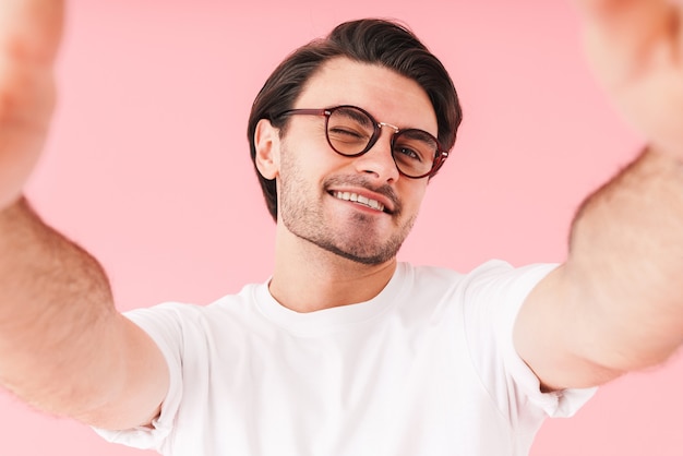 Obraz młodego radosnego mężczyzny noszącego okulary, mrugającego i robiącego zdjęcie selfie na białym tle