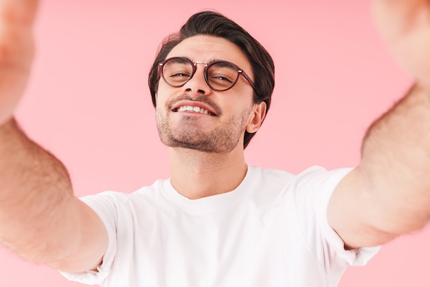 Obraz młodego radosnego mężczyzny noszącego okulary, uśmiechającego się i robiącego zdjęcie selfie na białym tle