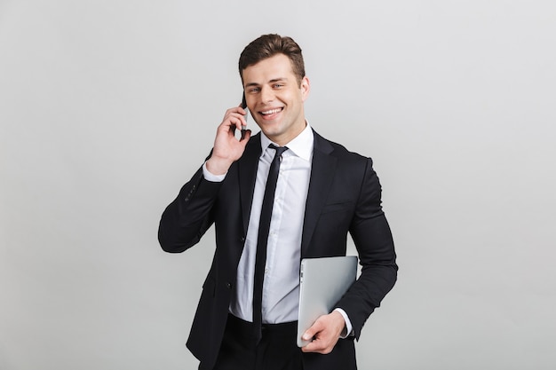 Obraz młodego uśmiechniętego biznesmena w formalnym garniturze rozmawiającego przez telefon komórkowy, trzymając laptopa na białym tle