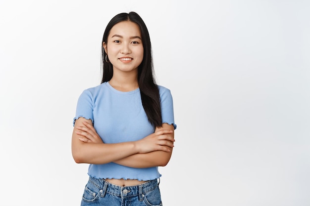 Obraz młodej azjatyckiej modelki krzyżującej się z ramionami na klatce piersiowej, która wygląda pewnie i uśmiecha się do kamery stojącej w zwykłych ubraniach na białym tle