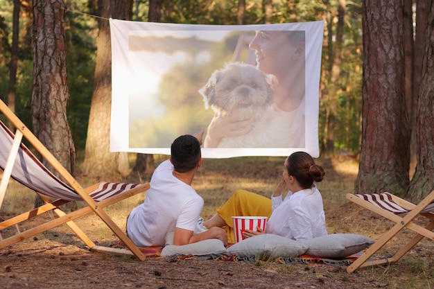 Obraz młodej pary leżącej w lesie z rzutnikiem i oglądającej film lub zdjęcia jedzącej popcorn w swobodnym stroju, cieszącej się ciekawym filmem i smaczną przekąską
