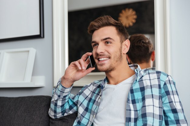 Obraz młody szczęśliwy człowiek ubrany w koszulę w klatce wydruku siedzi na kanapie w domu i rozmawia przez telefon.