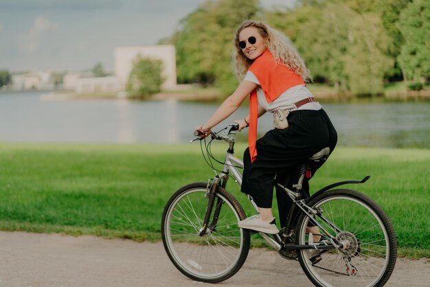 Obraz modelki jadącej na rowerze odwraca wzrok z radosnym wyrazem twarzy nosi okulary przeciwsłoneczne oddycha świeżym powietrzem pozy w pobliżu jeziora i zielonych drzew pokonuje duże odległości dowiaduje się czegoś nowego