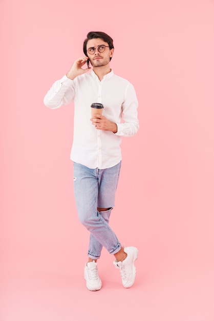Obraz nieogolonego, zakłopotanego mężczyzny w okularach, pijącego kawę na wynos i rozmawiającego przez telefon komórkowy na białym tle