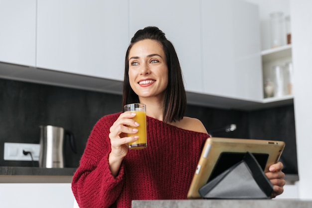 Obraz niesamowitej kobiety w domu w kuchni przy użyciu komputera typu tablet do picia soku.