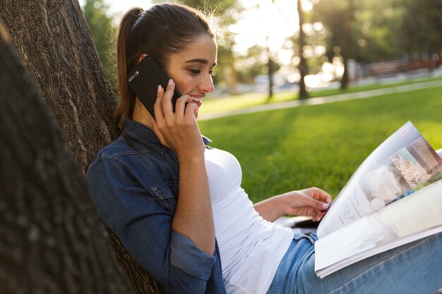 Obraz niesamowitej pięknej młodej kobiety studentki w parku trzymając zeszyt rozmawia przez telefon.