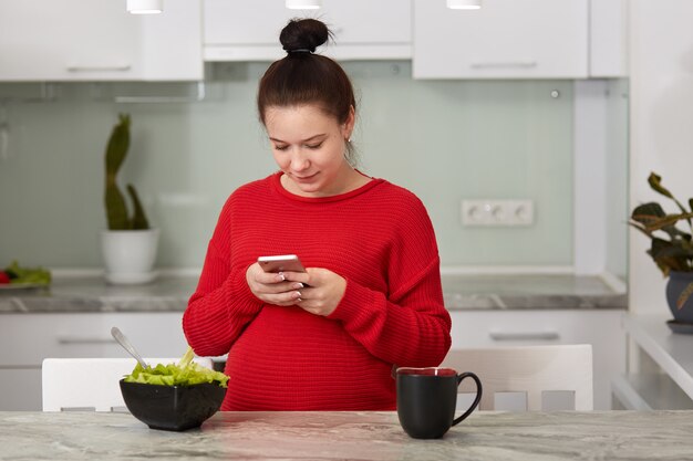 Obraz niesamowitej zdrowej kobiety w ciąży w kuchni w domu przy użyciu telefonu komórkowego do sprawdzania sieci społecznościowej lub poczty e-mail, jedzenia sałatki i picia herbaty lub kawy.