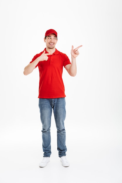 Obraz pełnej długości młodego kuriera w czerwonym mundurze przedstawiający tekst lub produkt copyspace z palcami wskazującymi na bok i szeroko uśmiechniętymi, odizolowane na białym tle