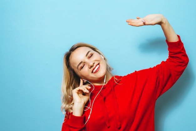 Obraz pięknej blondynki podekscytowana dziewczyna słucha muzyki w słuchawkach i tańczy na niebieską ścianą