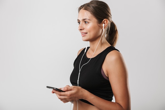 Obraz pięknej kobiety w odzieży sportowej trzymającej smartfon i słuchającej muzyki za pomocą słuchawek izolowanych na białej ścianie