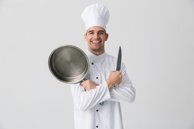 Obraz podekscytowany emocjonalny młody człowiek kucharz w pomieszczeniu na białym tle na tle białej ściany trzymając patelnię.