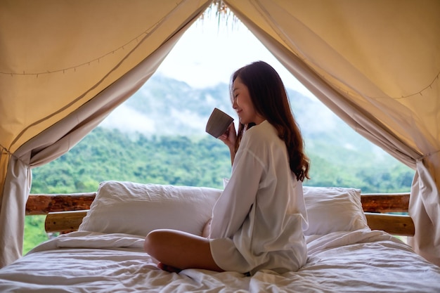 Obraz portretowy pięknej młodej kobiety azjatyckiej pijącej kawę siedząc na białym łóżku rano z pięknym widokiem na przyrodę poza namiotem