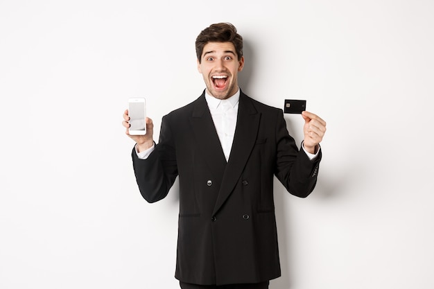 Obraz przystojnego biznesmena w czarnym garniturze, wyglądającego na podekscytowanego i pokazującego kartę kredytową z ekranem telefonu komórkowego, stojącego na białym tle
