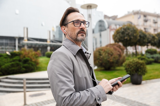 Obraz przystojnego biznesmena w okularach przy użyciu telefonu komórkowego i słuchawek podczas spaceru ulicą miasta