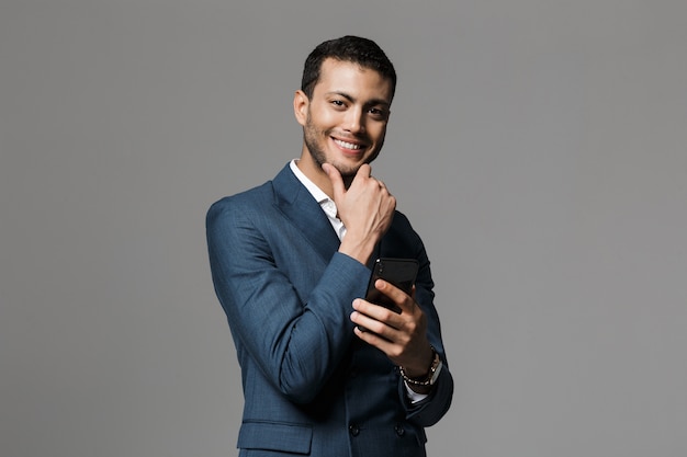 Obraz przystojny młody szczęśliwy wesoły biznesowy mężczyzna na białym tle na szarej ścianie za pomocą telefonu komórkowego.