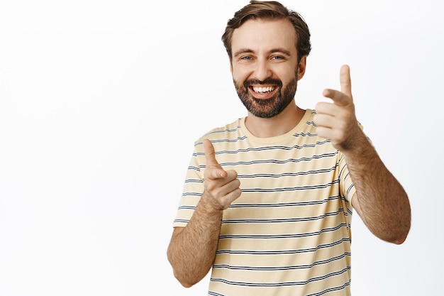 Obraz radosnego faceta z brodą wskazującego palcami na aparat i uśmiechniętego gratulującego chwalenia i komplementowania ciebie stojącego w koszulce na białym tle