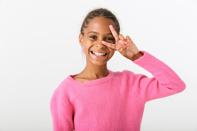 Obraz radosnej afroamerykańskiej dziewczyny uśmiechającej się i gestykulującej znak pokoju na białym tle nad białą ścianą