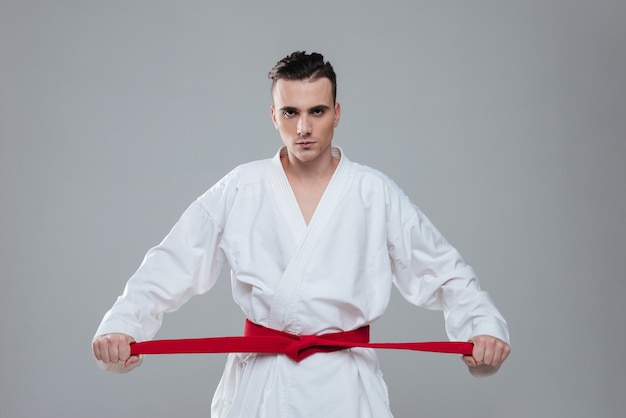 Obraz sportowca ubrany w praktykę kimono w karate podczas zaciskania czerwonego pasa na białym tle nad szarym tłem. Spójrz na aparat.