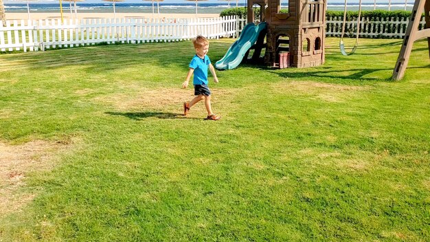 Obraz szczęśliwego uśmiechniętego i roześmianego chłopca biegającego po zielonej trawie na placu zabaw dla dzieci