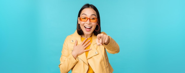 Obraz szczęśliwej koreańskiej kobiety w okularach przeciwsłonecznych wskazujący palec na aparat z wyrazem twarzy zdumiony, zaskoczony i radosny, stojący na niebieskim tle