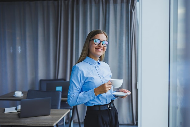 Obraz szczęśliwej młodej kobiety w klasycznej koszuli i okularach uśmiecha się i pije kawę stojąc przy oknie w nowoczesnym biurze z dużymi oknami Praca zdalna