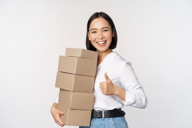 Obraz szczęśliwej nowoczesnej azjatyckiej kobiety pokazującej kciuki do góry trzymającej pudełka z towarami dostawczymi stojące na białym tle