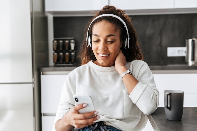 Obraz szczęśliwy African American girl noszenia słuchawek za pomocą smartfona, siedząc w jasnej kuchni