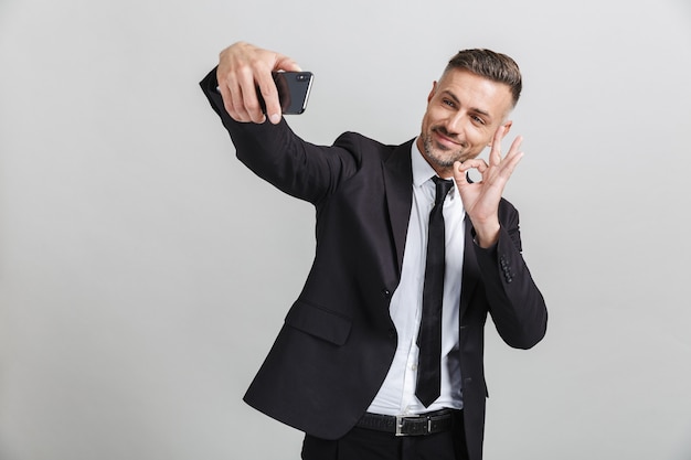 Obraz udanego uśmiechniętego biznesmena w formalnym garniturze, gestykulującego ok śpiewać podczas robienia selfie na telefonie komórkowym na białym tle