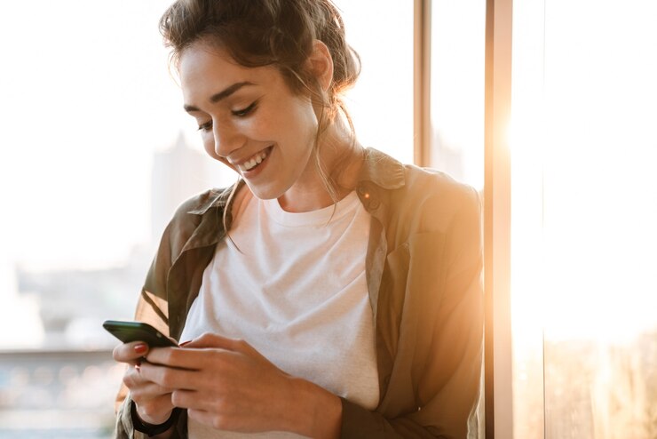 Zdjęcie obraz uroczej brunetki uśmiechającej się i trzymającej smartfon stojąc na tarasie na zewnątrz rano