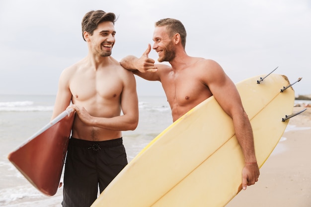 Obraz uśmiechający się wesoły dwóch mężczyzn, którzy surferów uprawiają surfing na plaży na zewnątrz rozmawiając ze sobą.