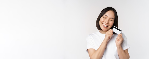 Obraz uśmiechniętej azjatyckiej kobiety przytulającej kartę kredytową, kupującej zbliżeniowo, stojącej w białej koszulce na białym tle