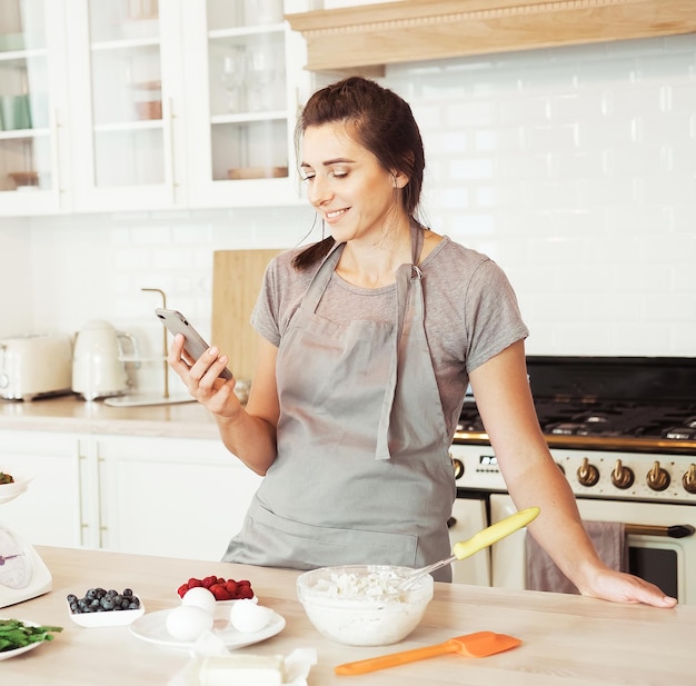 Obraz uśmiechniętej kobiety w fartuchu przy użyciu telefonu komórkowego podczas gotowania ciasta w nowoczesnej kuchni
