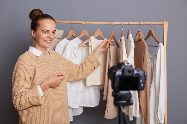 Obraz uśmiechniętej ładnej kaukaskiej blogerki pozującej w szafie stojącej przed wieszakiem ze strojami nagrywa wideo sprzedające jej ubrania online