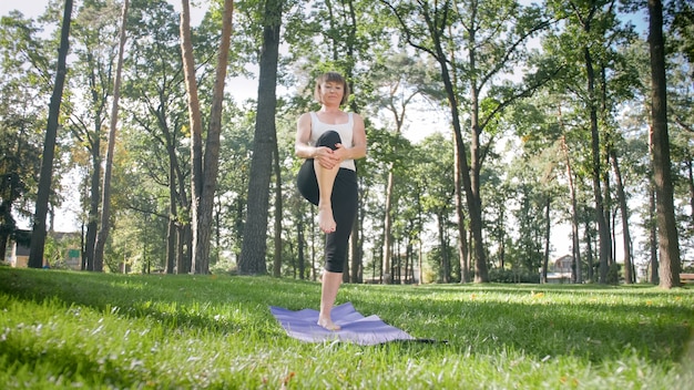 Obraz w średnim wieku uśmiechnięta szczęśliwa kobieta medytuje i robi ćwiczenia jogi na trawie w lesie. Kobieta dbająca o zdrowie fizyczne i psychiczne podczas ćwiczeń fitness i rozciągania w parku