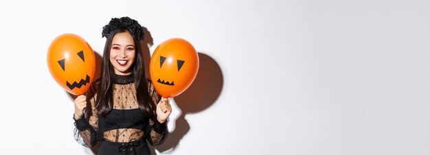 Obraz wesołej azjatyckiej kobiety w stroju wiedźmy świętującej halloween, trzymającej balony z przerażającą twarzą