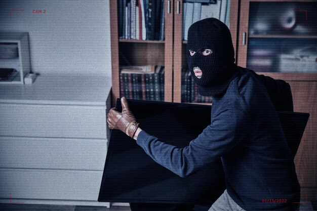 Obraz z kamery bezpieczeństwa ze złodziejem z czarną kominiarką i ciemnymi ubraniami kradnącymi telewizor