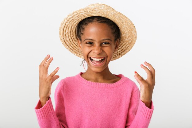 Obraz zachwyconej afroamerykańskiej dziewczyny w słomkowym kapeluszu, śmiejącej się z uniesionymi rękami, odizolowanej na białej ścianie