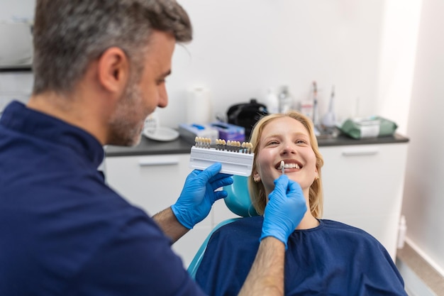 Obraz zadowolonej młodej kobiety siedzącej na fotelu dentystycznym w centrum medycznym, podczas gdy profesjonalny lekarz naprawia jej zęby Dentysta wybiera odcień wypełnienia dla uśmiechniętej kobiety za pomocą próbki łusek zębów