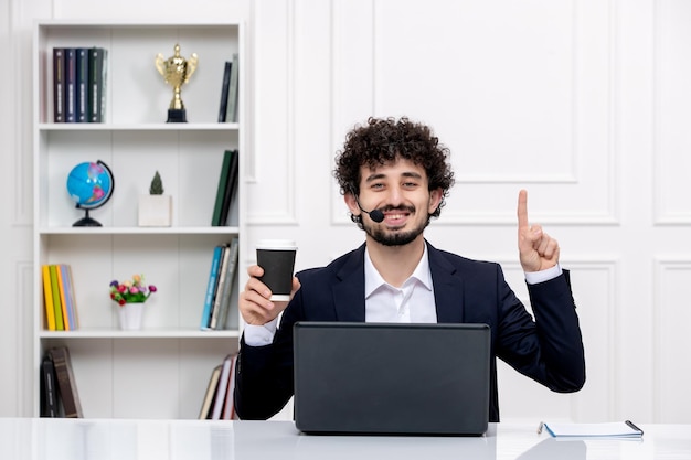 Obsługa klienta przystojny kędzierzawy mężczyzna w garniturze biurowym z komputerem i zestawem słuchawkowym zadowolony z filiżanki kawy