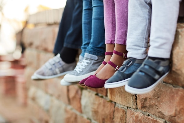 Obuwia nogi dzieci w wieku szkolnym i siedzenie razem na ceglanym murze Przyjaźń grupy przyjaciół i młody student poza salą lekcyjną w porze lunchu lub przerwy z pokazanym obuwiem