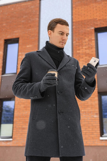 Od dołu skoncentrowanego męskiego przedsiębiorcy w zimowym płaszczu z filiżanką kawy za pomocą smartfona
