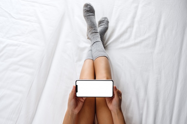 Odgórny Widok Kobieta Trzyma Białego Telefon Komórkowego Z Pustym Desktop Ekranem Podczas Gdy Kłamający Na Wygodnym Białym łóżku W Domu