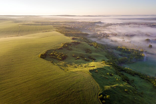 Odgórny widok zieleń kultywujący wzgórza, zieleni drzewa i mali domy w mgłowej dolinie na jaskrawej niebieskie niebo kopii przestrzeni. Wiosny mglista krajobrazowa panorama przy świtem.