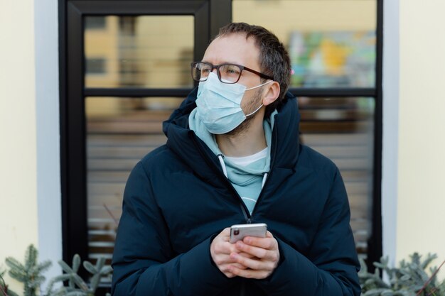 Odkryty strzał zadumanego mężczyzny nosi maskę ochronną przed koronawirusem na ulicy, trzyma telefon komórkowy, czyta wiadomości, dba o zdrowie, korzysta z bezprzewodowego internetu. Opieka zdrowotna. Covid-19. Wirus rozprzestrzenia się